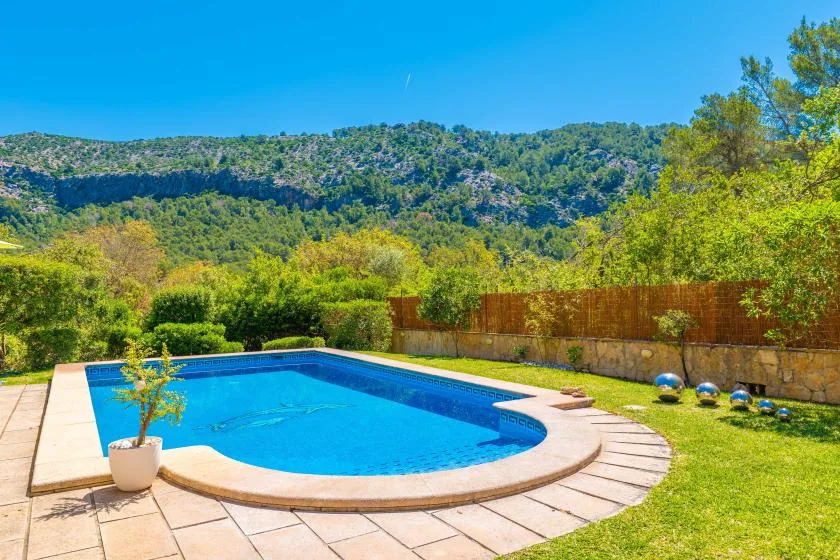 An outdoor swimming pool sits beside a paved patio, with wooded hills beyond.