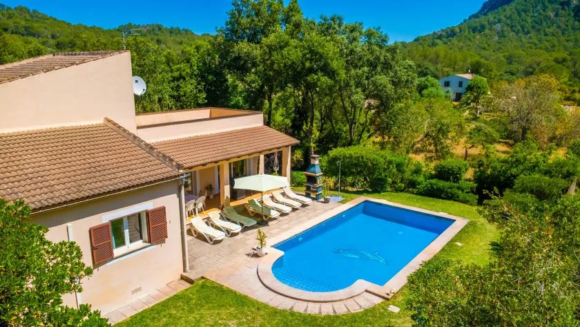 A house and bright blue swimming pool sit amid green trees and hills.