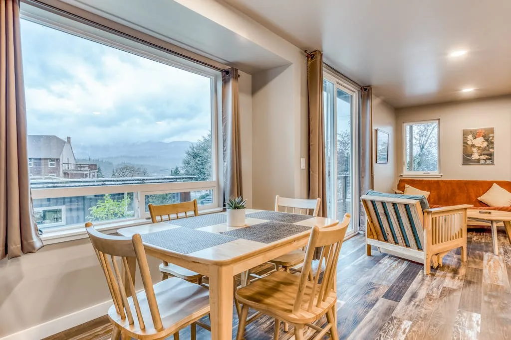 Zona de comedor y estar con una mesa de madera, sillas y ventanales.