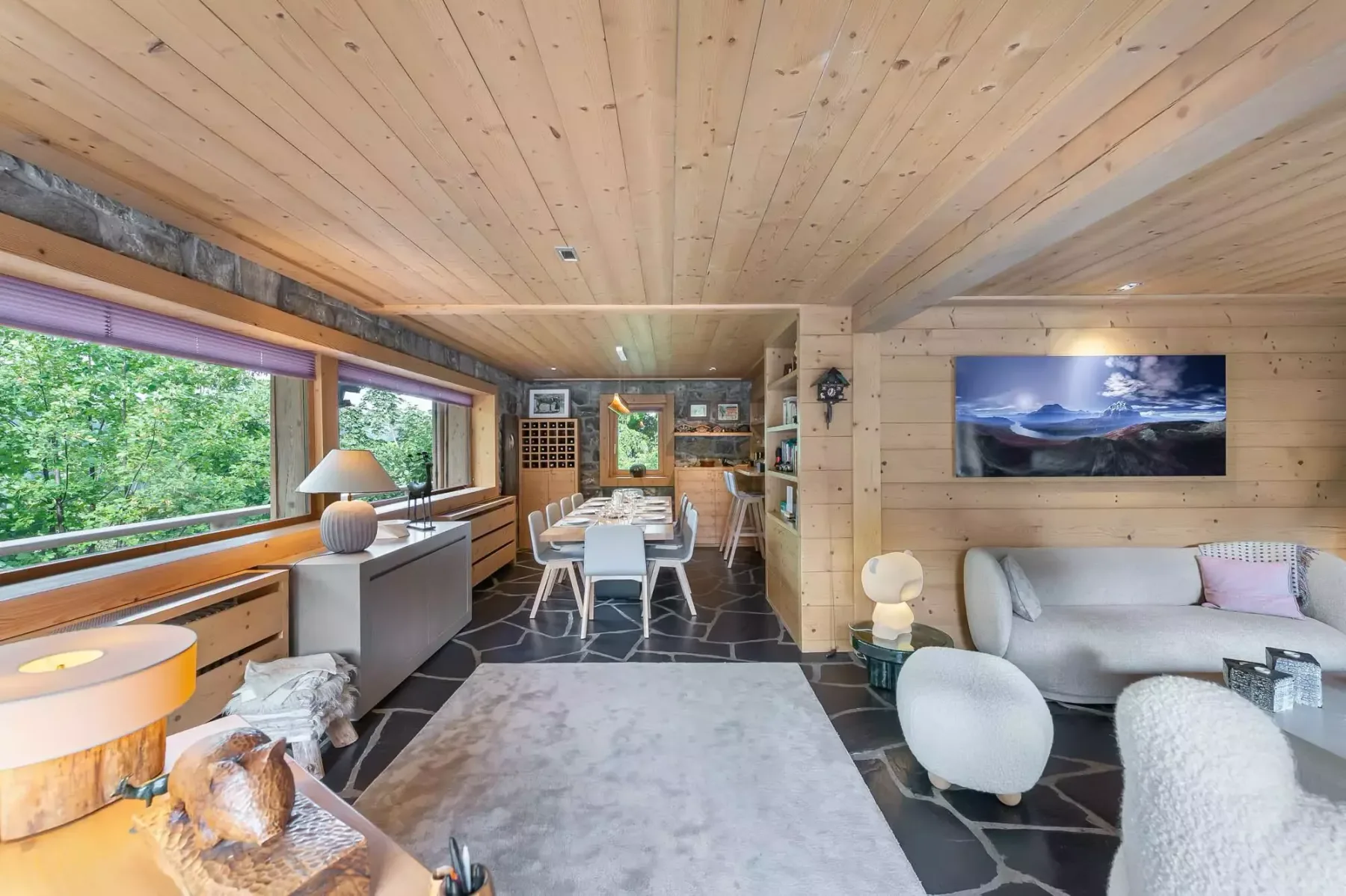 Sala de estar y comedor revestidos de madera, con mesa, asientos y grandes ventanas.