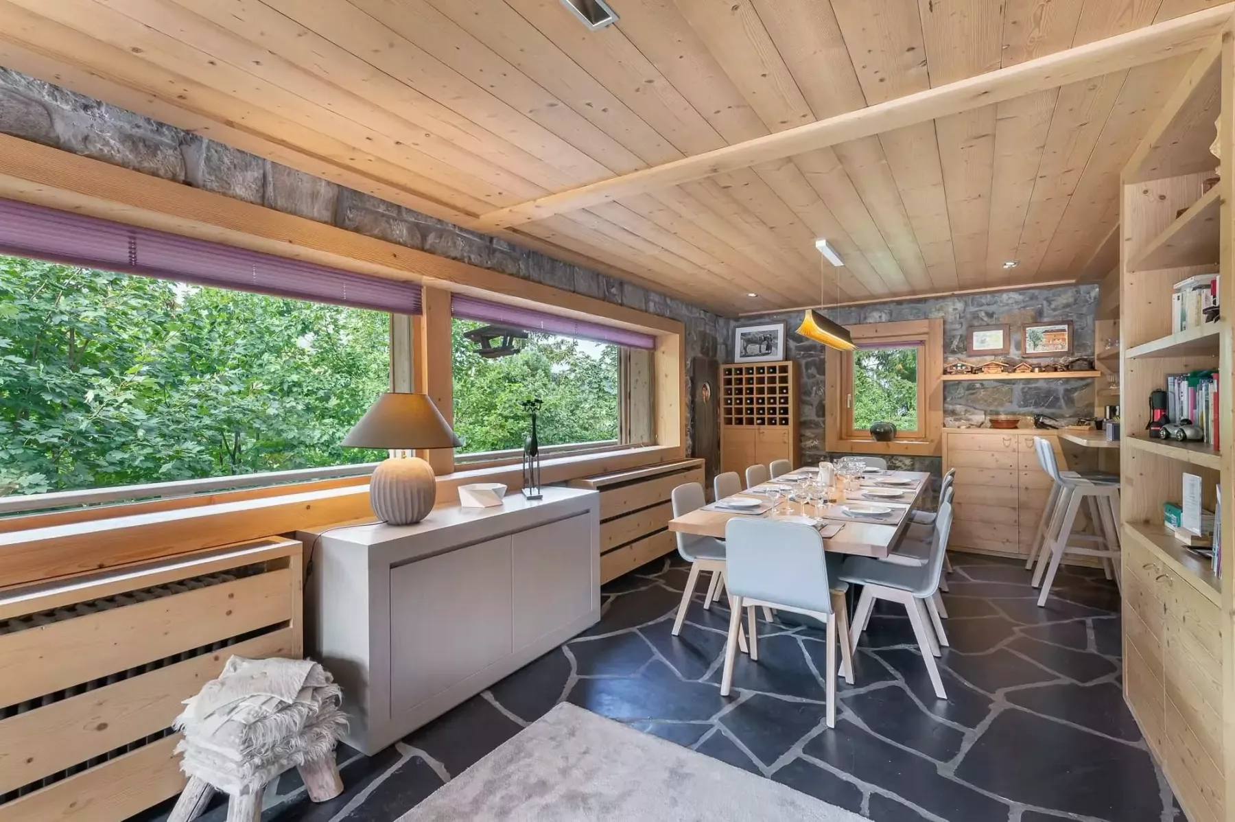 A dining area with a long table beside large windows overlooking greenery.