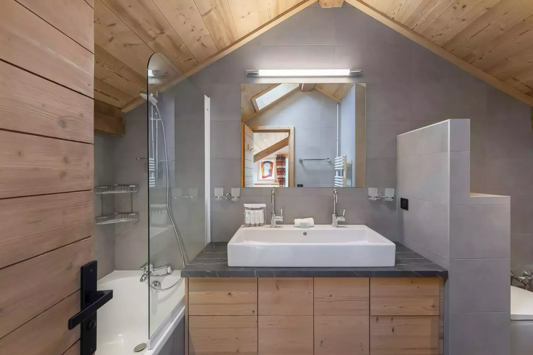 Un baño con bañera, mampara de ducha de vidrio y un amplio lavabo bajo un techo inclinado.