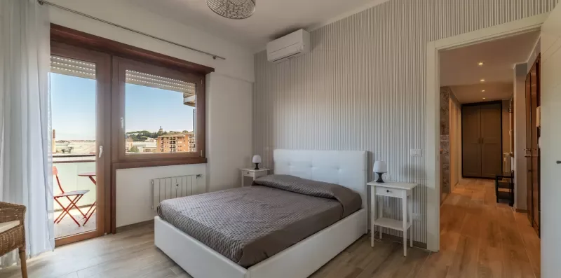 Un dormitorio con una cama doble junto a una ventana amplia con vistas a edificios cercanos.