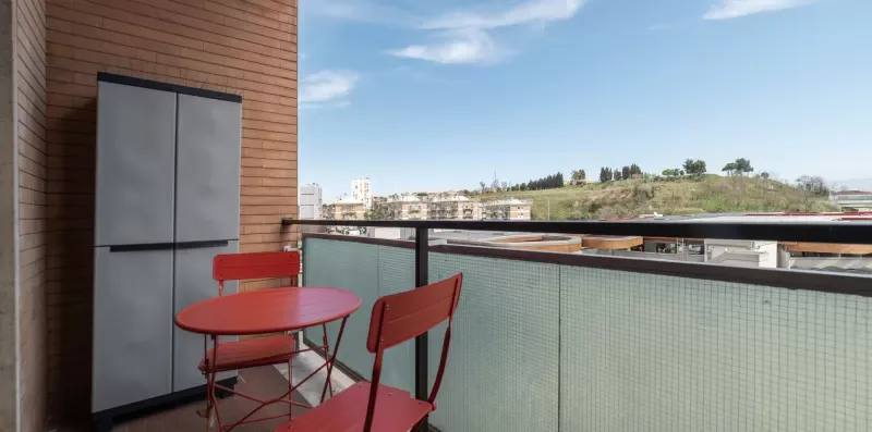 Un balcón con asientos rojos ofrece vistas de edificios cercanos y una colina verde.