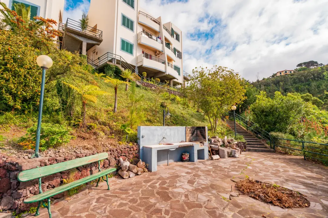 A paved outdoor terrace with a built-in barbecue, benches, and steps beside a landscaped slope.