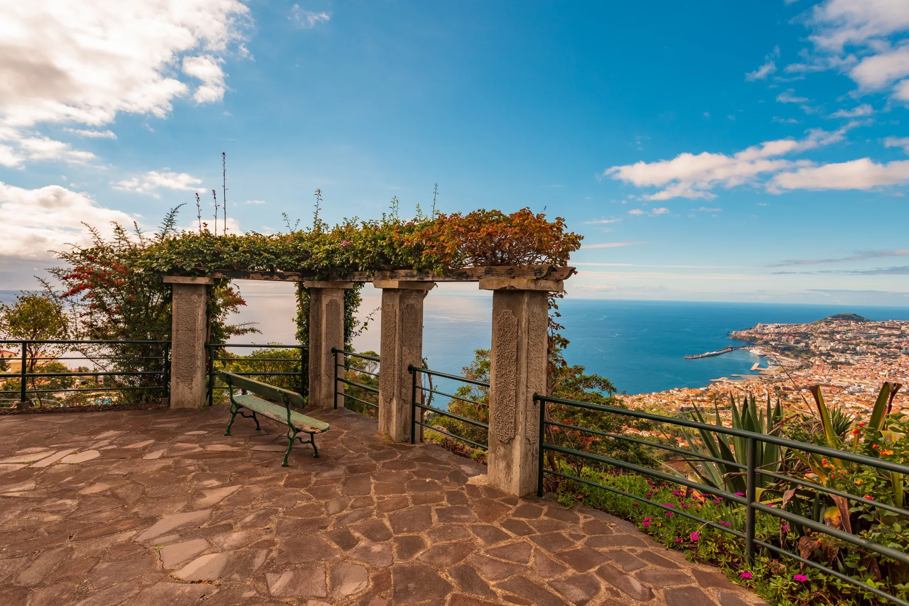 A stone-paved terrace overlooks the ocean and a coastal city.