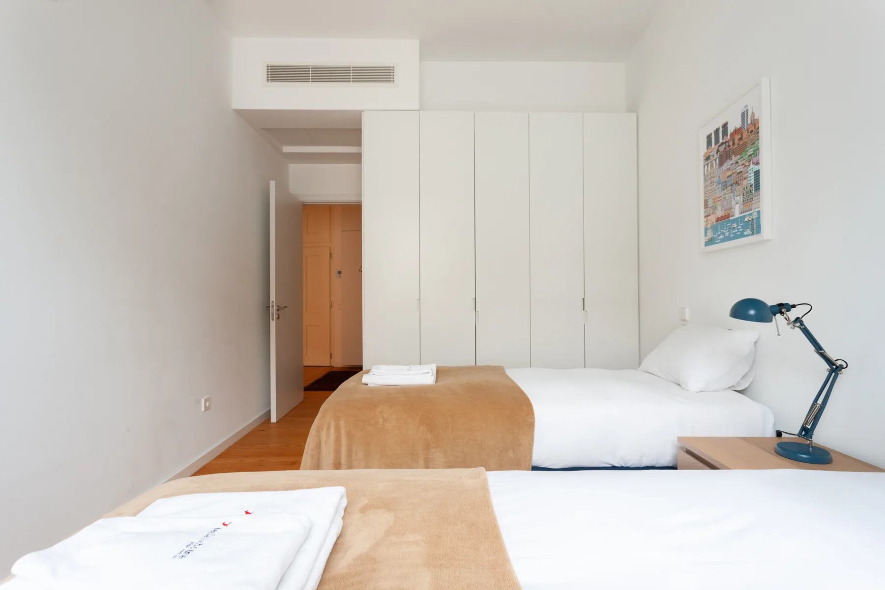 Un dormitorio luminoso con dos camas, toallas dobladas y una lámpara de escritorio.