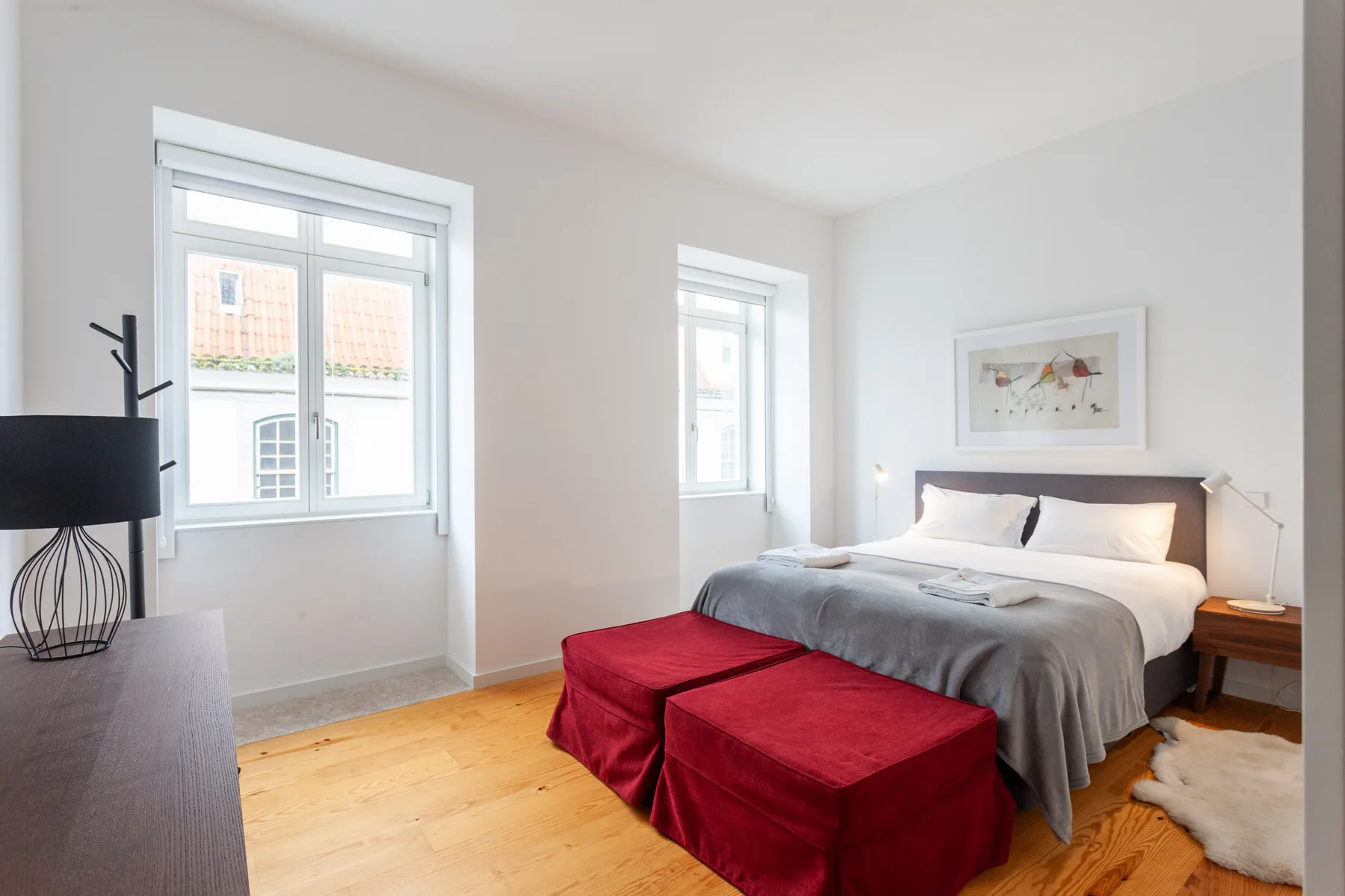 Un dormitorio luminoso con una cama grande, pufs rojos y dos ventanas.