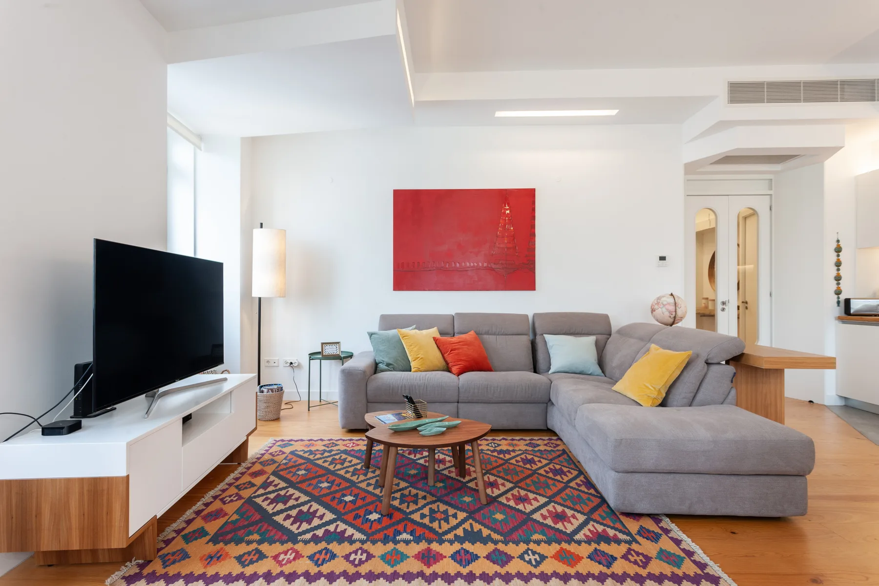 Una sala de estar luminosa con asientos grises, un televisor y una alfombra estampada colorida.
