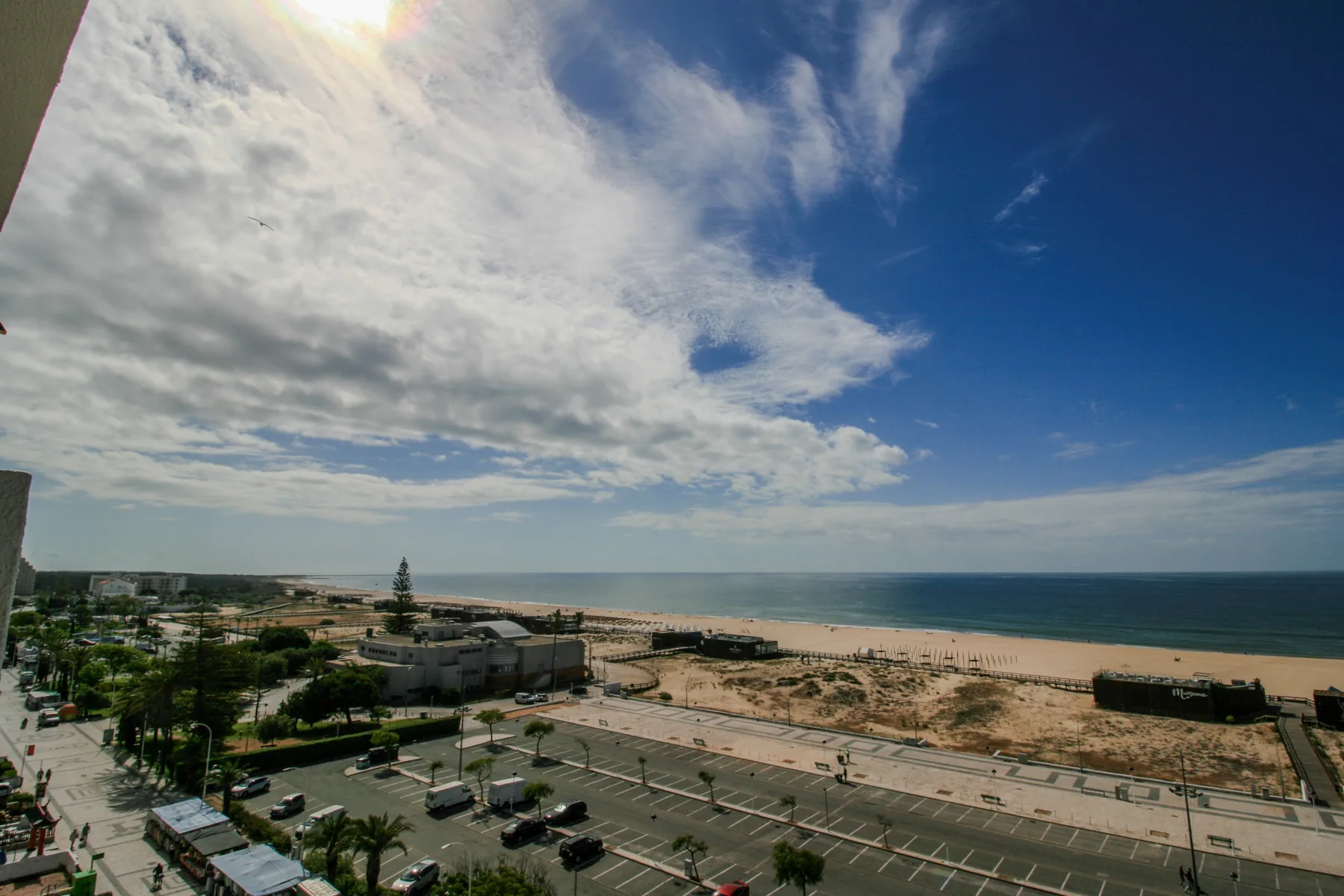 A coastal view overlooks the beach, ocean, and a parking area beneath a cloudy sky.