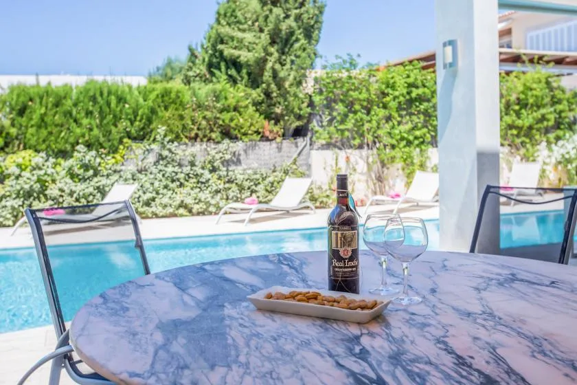 A patio table overlooks a swimming pool, with wine glasses and a bottle.