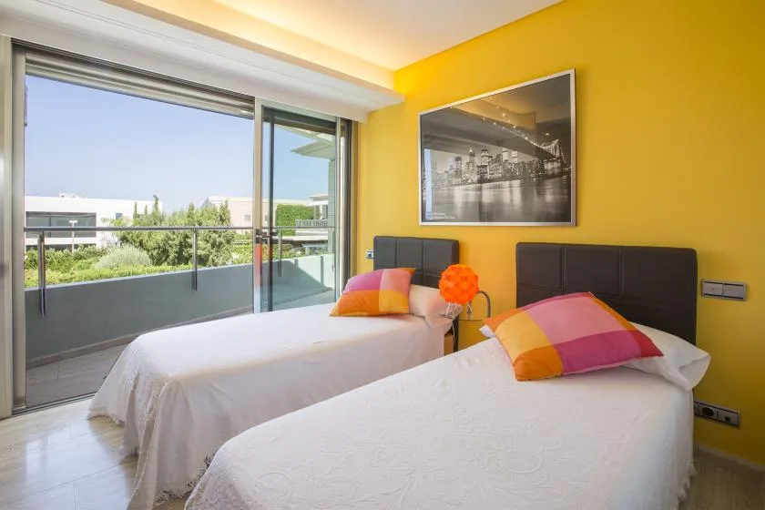 Un dormitorio luminoso con dos camas junto a una gran puerta acristalada al balcón.