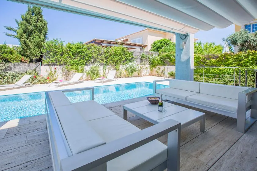 A covered poolside seating area overlooks a swimming pool and greenery.