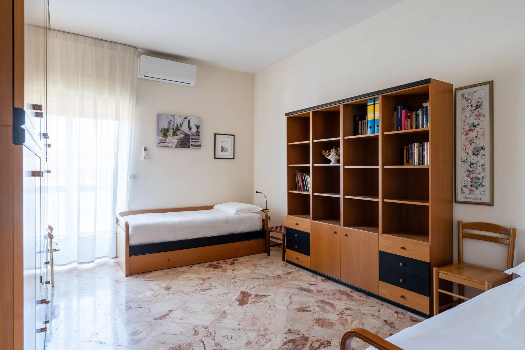 A bedroom with a single bed, a tall bookcase, and a bright window.