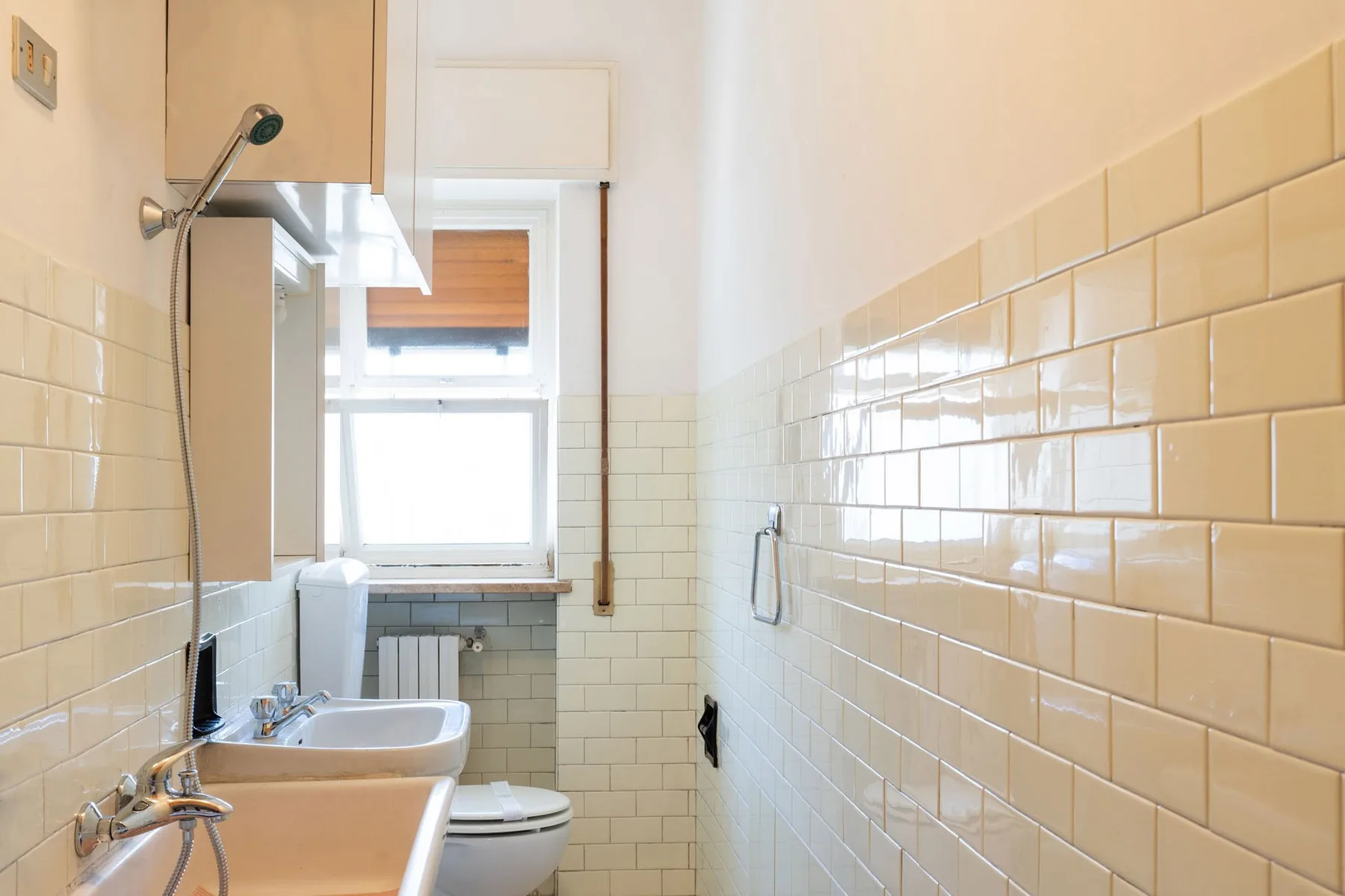 A narrow tiled bathroom with a shower, sink, toilet, and window.