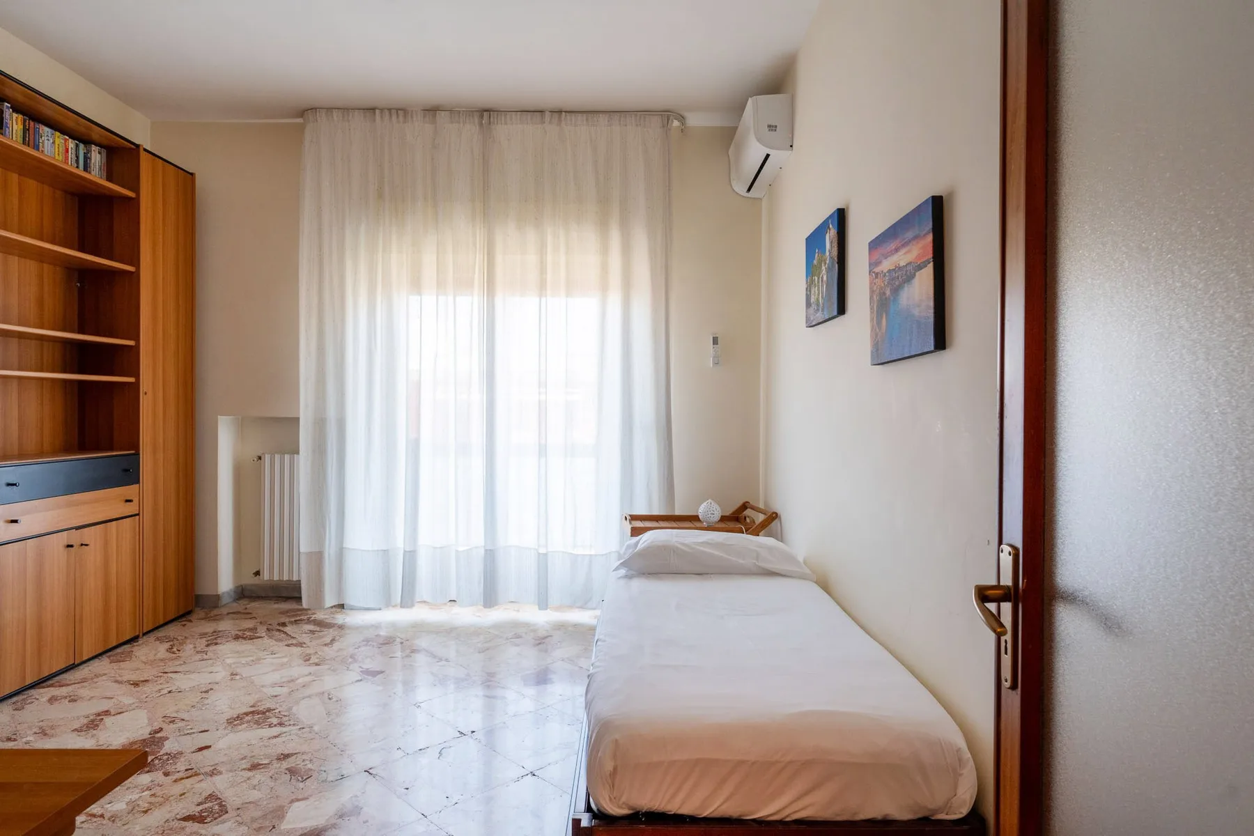 A bright bedroom with a single bed, sheer curtains, and wooden shelving.