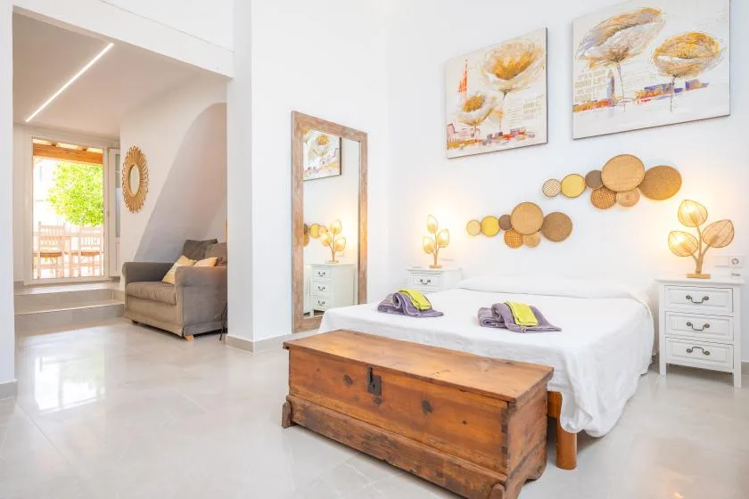 Un dormitorio luminoso con una cama, un baúl de madera y cuadros en la pared.