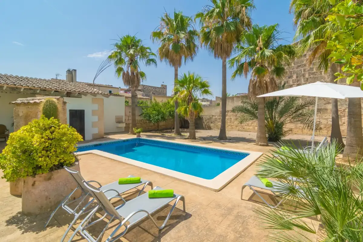 Una zona de piscina al aire libre, soleada, con palmeras, asientos reclinables y una sombrilla.