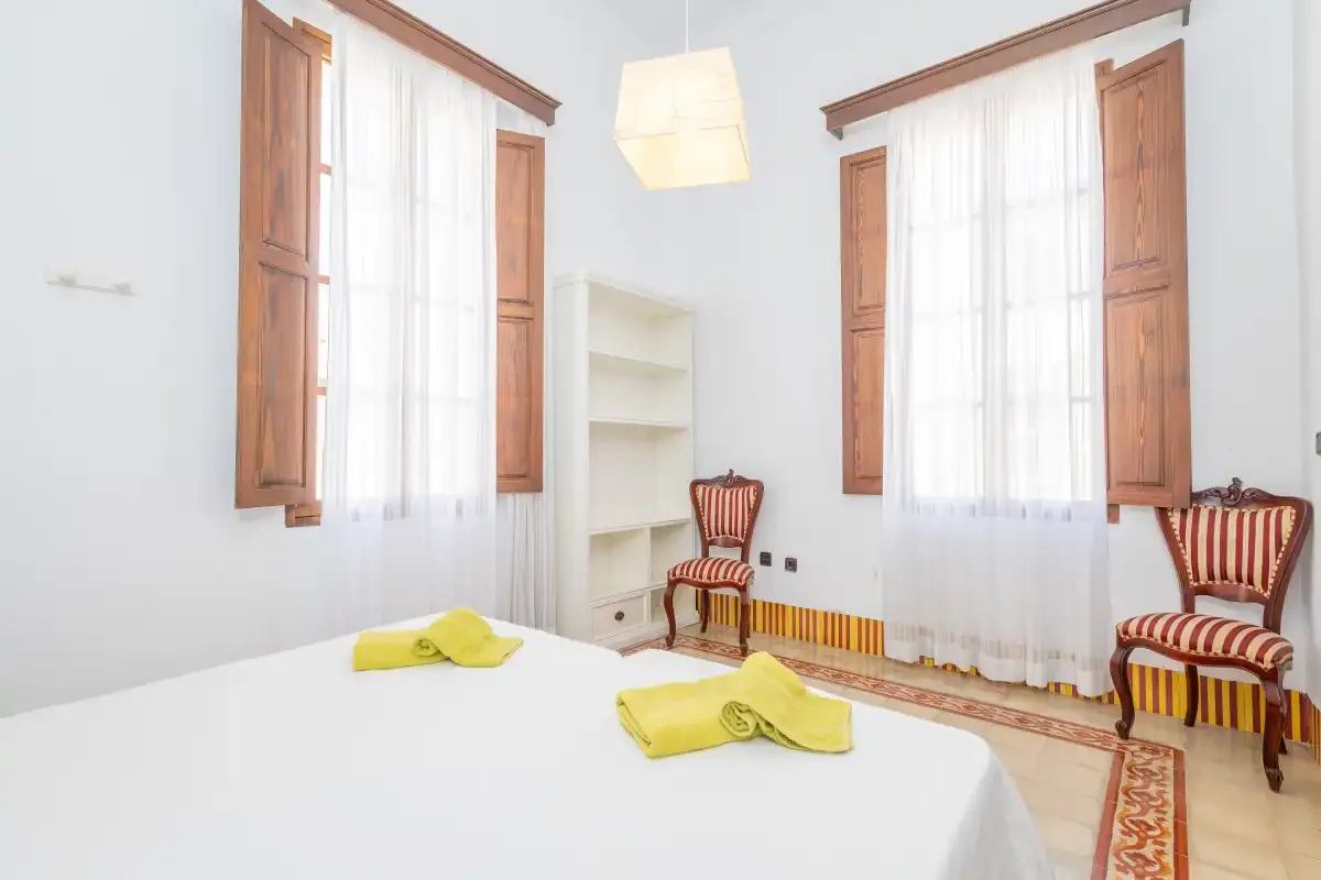Un dormitorio luminoso con una cama, ventanas altas y asientos a rayas.