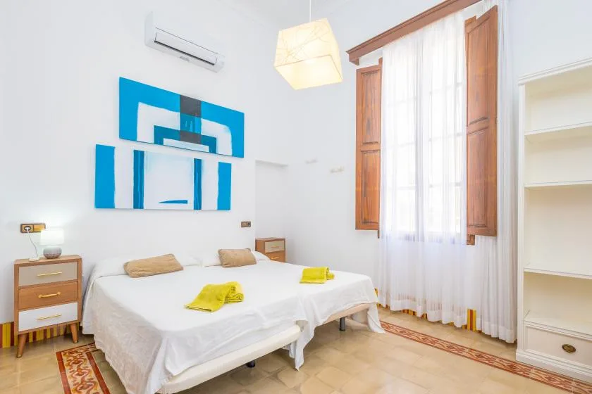 Un dormitorio con una cama, arte mural azul y una ventana alta con cortinas.