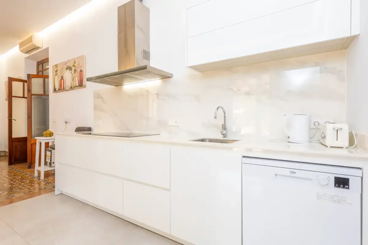 Una cocina blanca y luminosa con encimera, fregadero y pequeños electrodomésticos.