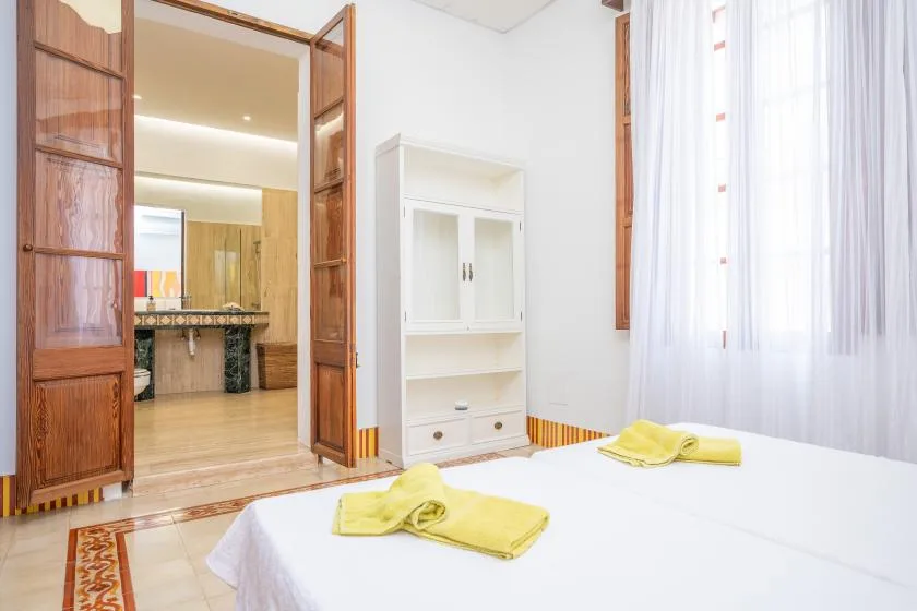 Un dormitorio con una cama blanca y puertas de madera que dan a una estancia con suelo de baldosas.