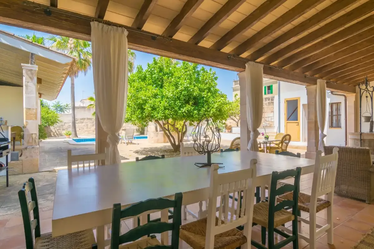 Una zona de comedor exterior cubierta se abre a un jardín con un árbol frondoso.