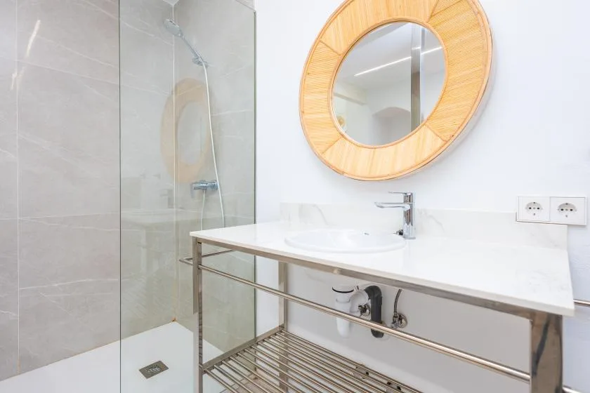 Un baño luminoso con una ducha de vidrio y un espejo redondo sobre el lavabo.