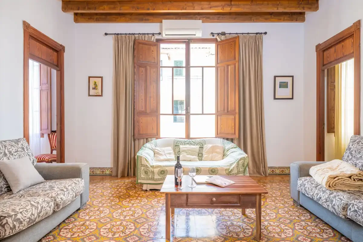Una sala luminosa con asientos, una mesa central y ventanas de madera abiertas.