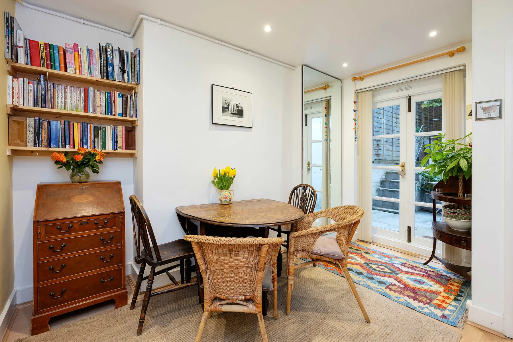 A bright dining area with a round wooden table, wicker seating, and a bookcase.
