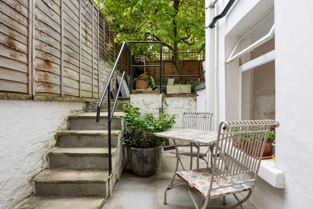 A small outdoor patio with a table, seating, and steps beside a leafy garden.