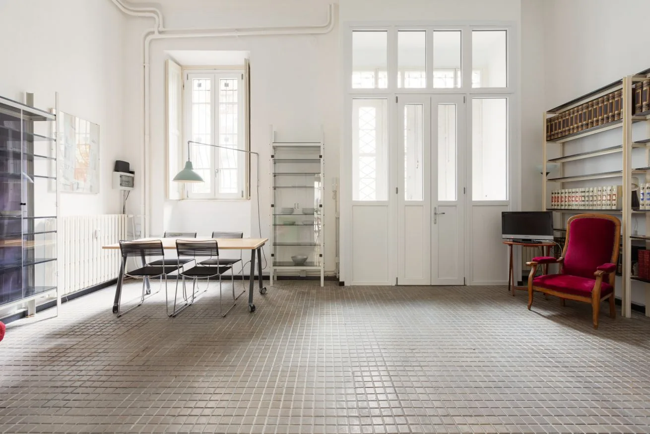 Una habitación luminosa con una mesa y asientos junto a ventanas altas y puertas de vidrio.