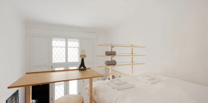 Un dormitorio luminoso con una cama blanca y un escritorio de madera junto a la ventana.