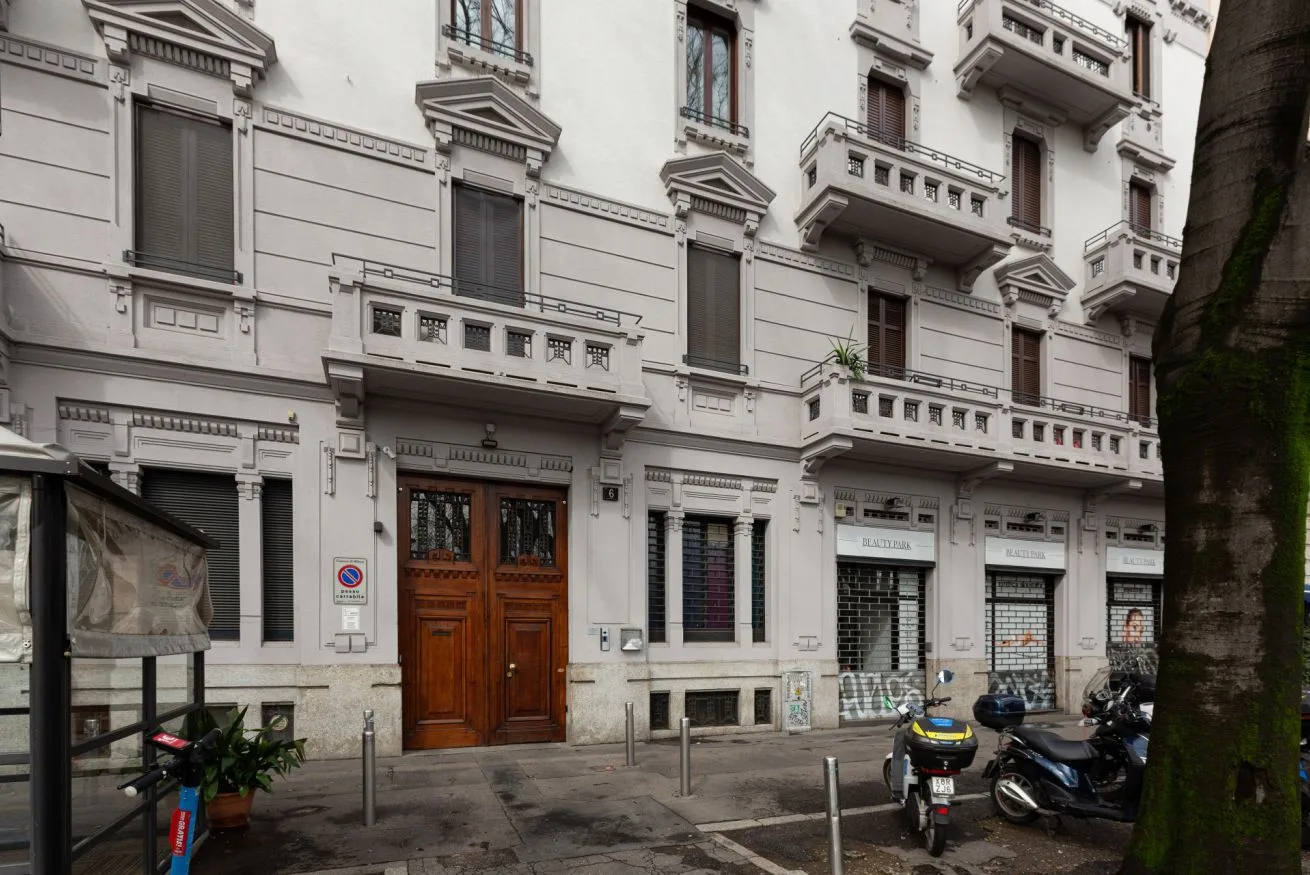 Fachada de un edificio blanco de varias plantas con balcones, locales y motos estacionadas.