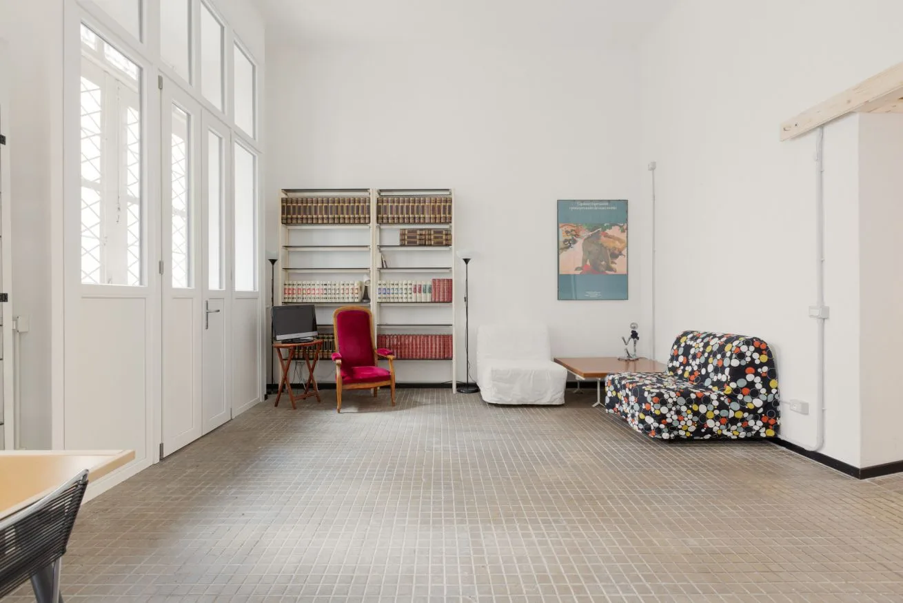 Una habitación luminosa con ventanas altas, estanterías y una pequeña zona de asientos.