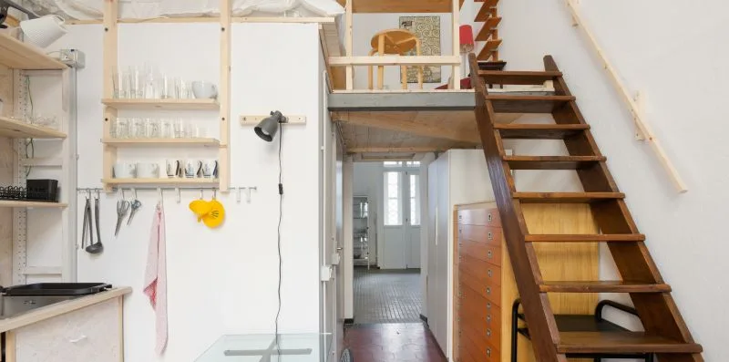Una pequeña zona de cocina está bajo una escalera de madera y una plataforma elevada.
