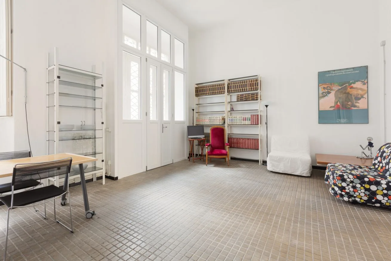 Una habitación luminosa con ventanas altas, estanterías y una pequeña zona de asientos.