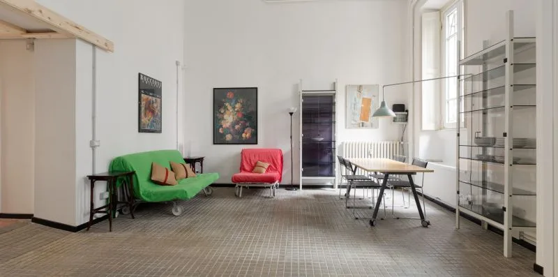 Una habitación luminosa con asientos verdes, una mesa, estanterías y ventanas grandes.