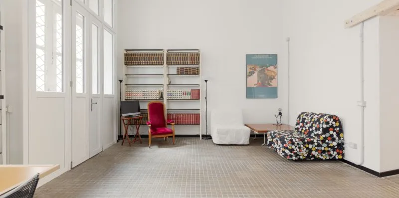 Una habitación luminosa con ventanas altas, estanterías y distintos asientos.