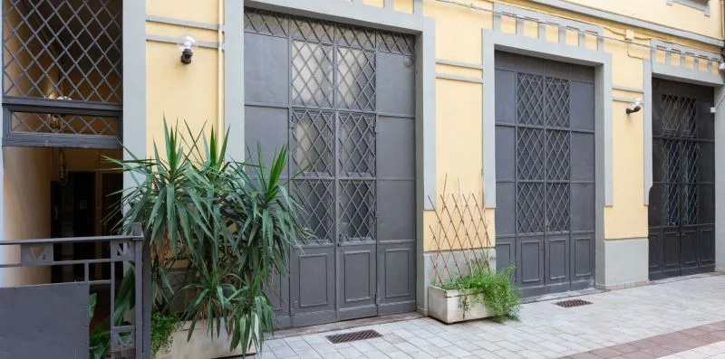 Una fachada amarilla con puertas metálicas oscuras y plantas en macetas junto a la acera.