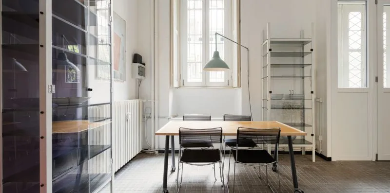 Un comedor luminoso con una mesa de madera, asientos y ventanas altas.