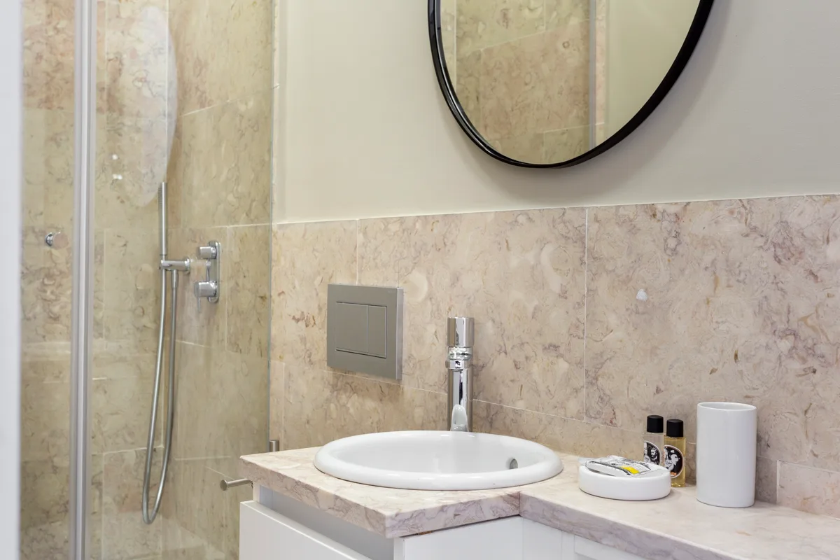 A bathroom vanity with a sink, toiletries, and a round mirror beside the shower.
