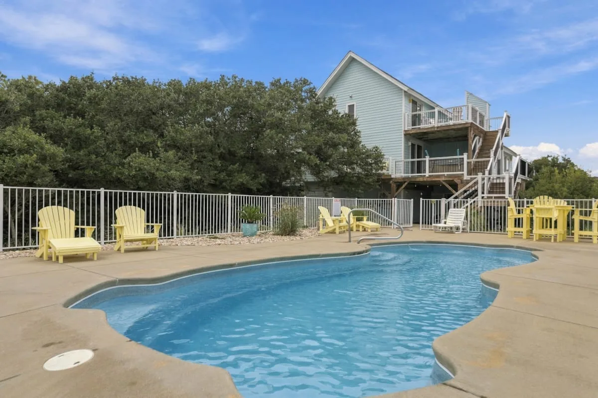 Una piscina al aire libre está junto a una terraza con asientos y una casa elevada.