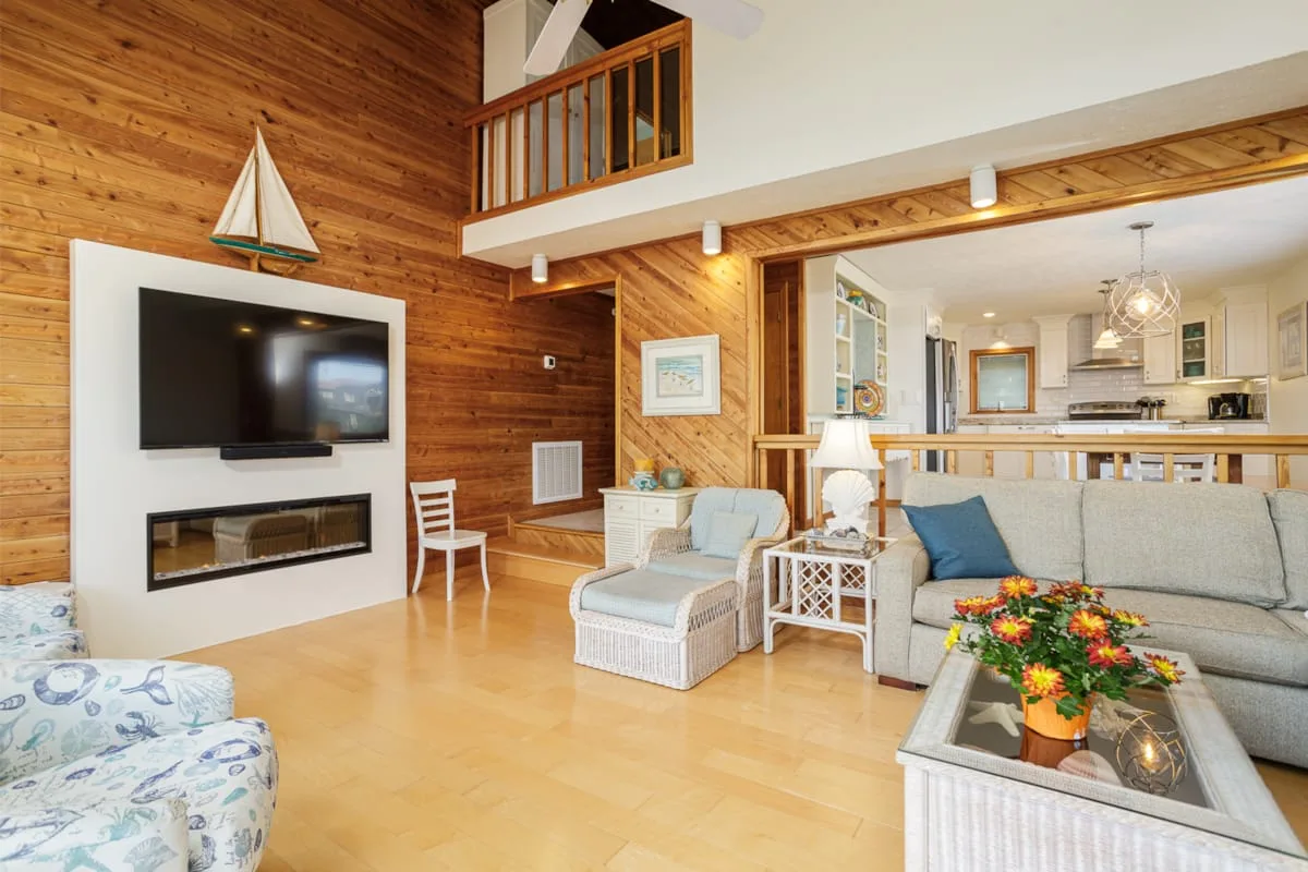Una sala de estar con paredes de madera se abre a una cocina y comedor iluminados.