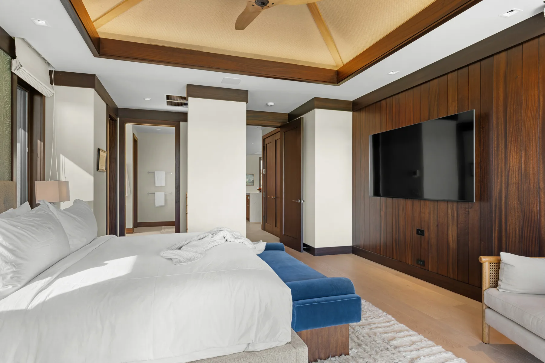 Un dormitorio con ropa de cama blanca, un banco azul y un televisor en la pared.
