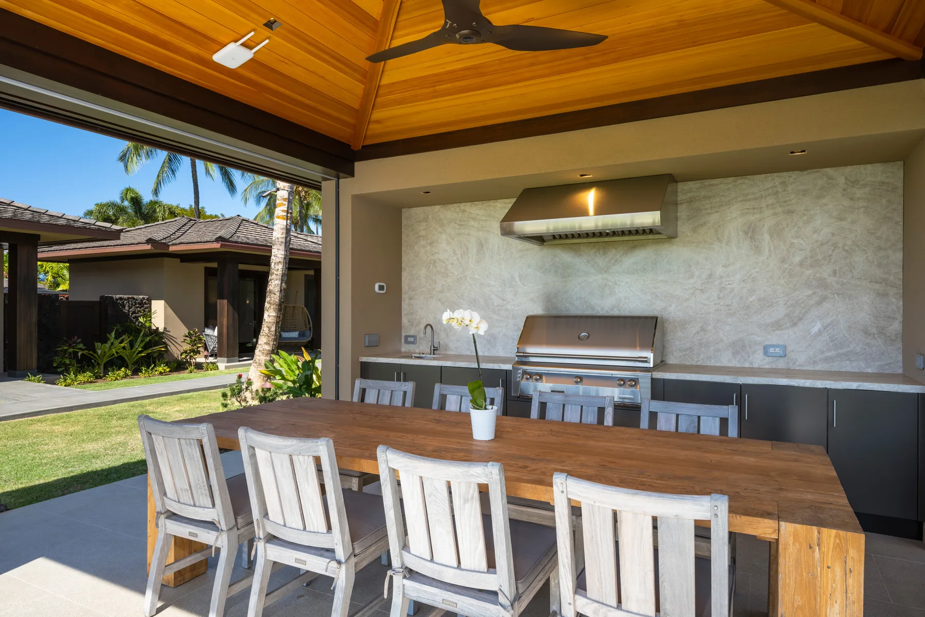 Comedor al aire libre con una mesa de madera bajo un patio cubierto y una parrilla empotrada.