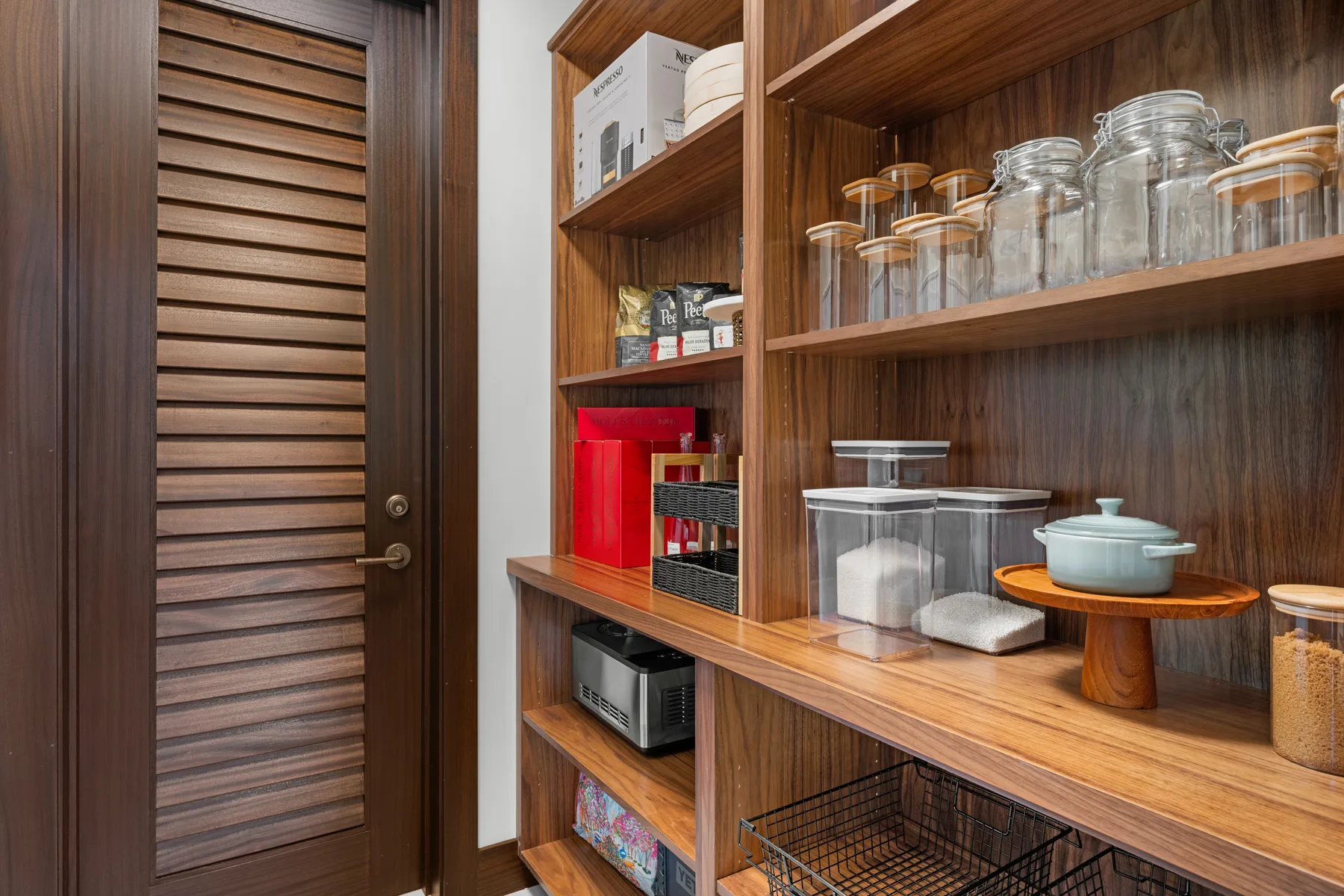 Una despensa de madera con estantes abiertos contiene frascos, recipientes y utensilios de cocina.