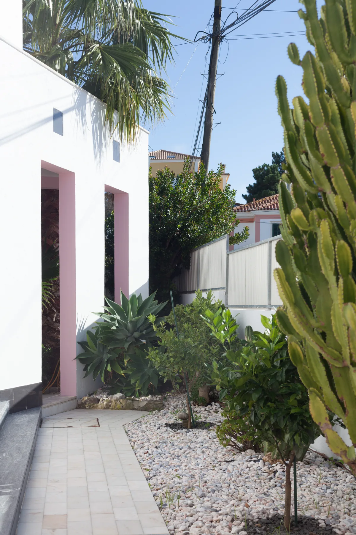 A paved garden path runs beside a white building and leafy plants.