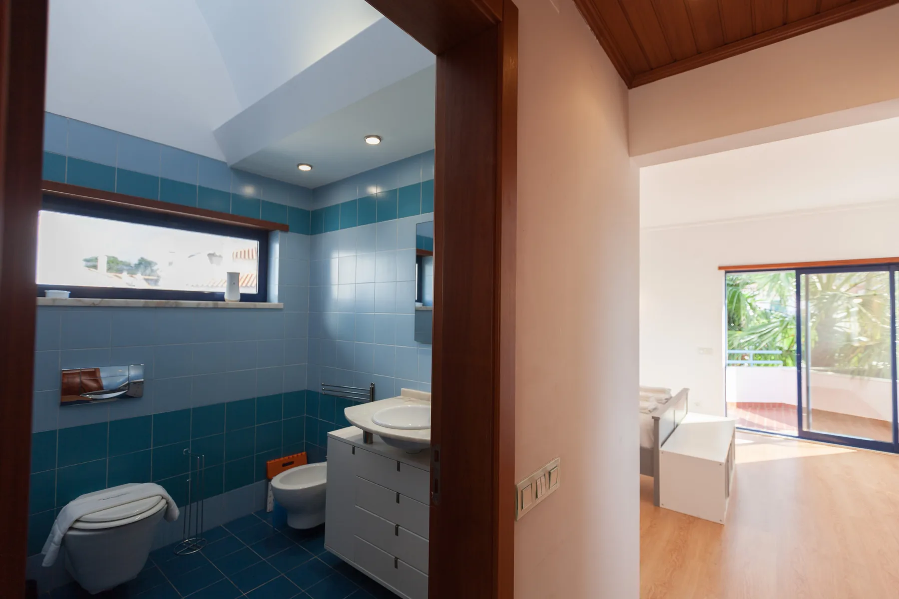 A blue-tiled bathroom with a toilet and sink opens onto a bright room.