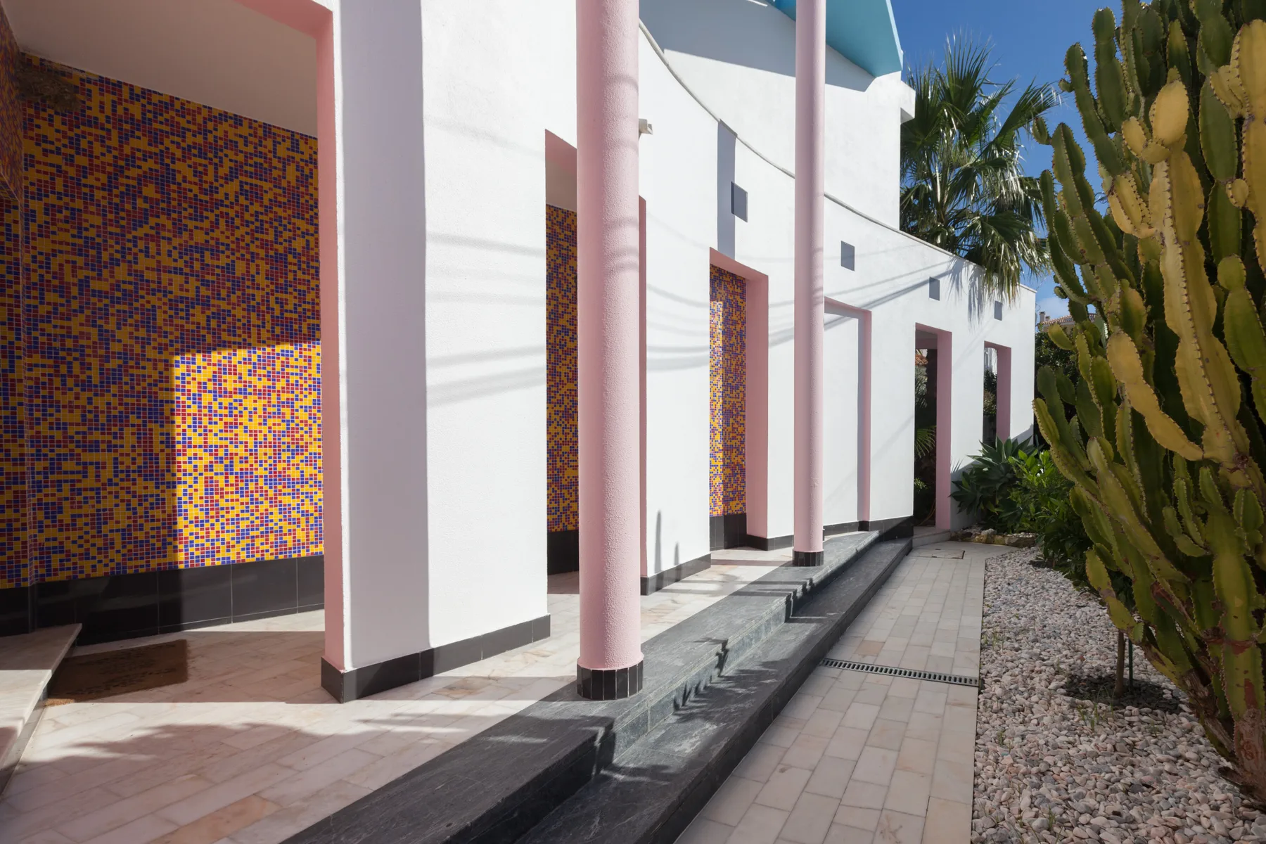 A paved outdoor walkway runs beside a white building with colorful tiled walls.